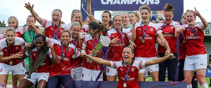 Arsenal WFC players celebrating with the league trophy under a Champions banner